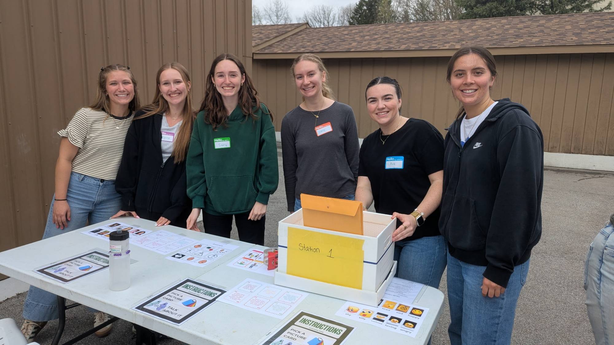 A group of six young adults standing behind a table with flyers and a labeled box outdoors.